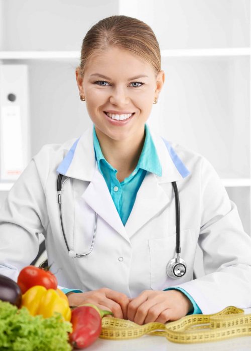 Happy smiling female dietitian in uniform with stethoscope at workplace. Young  woman doctor sitting at the desk with eco food.